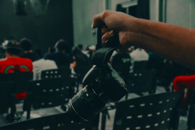A hand holding a camera by its strap in focus in the foreground, with a blurred background of people seated in rows of chairs, suggesting a possible event or gathering.