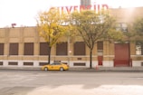 A yellow taxi is parked on a quiet street in front of a brick industrial building with a red rolled-up door and several rectangular windows. The building is partially obscured by two trees with yellowing leaves, indicating autumn. A large sign on the roofline hints at the building's function. The street is empty and a small orange traffic cone is placed on the sidewalk.