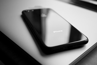 A sleek, black iPhone rests atop a closed MacBook. The surfaces reflect minimal light, showcasing their polished finish. The lighting highlights the phone's glossy texture and the distinct Apple logo on the laptop.