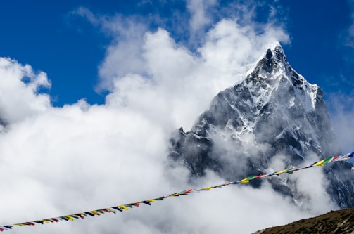 A majestic mountain peak covered in snow rises through the clouds. Colorful prayer flags are strung across the foreground, adding a vibrant contrast to the natural scene.