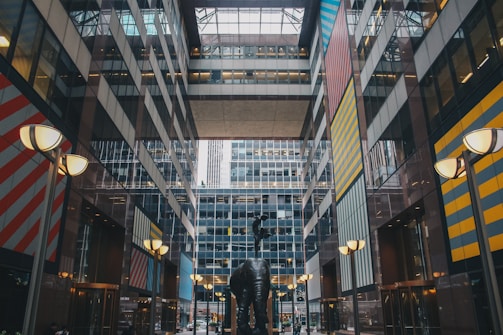 A modern architectural space features a striking abstract sculpture in the foreground, centered in a courtyard. The tall buildings surrounding have reflective glass facades and colorful geometric patterns on their walls, including stripes in red, blue, and yellow. The atmosphere is urban and dynamic.