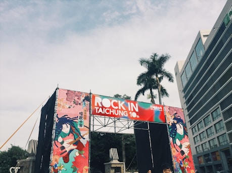A stage setup with a large banner displaying 'Rock in Taichung 10th' in bold white letters against a red and blue background. The sides of the stage feature artistic illustrations of musicians playing guitars. A tall palm tree is visible nearby, and an adjacent modern building with numerous windows can be seen.