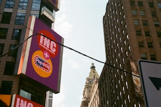 A cityscape featuring tall buildings with a prominent billboard displaying the logos for INC and Popsicle. A historical-style building with a detailed spire is visible in the background. The sky is partly cloudy, and a power line runs across the image.