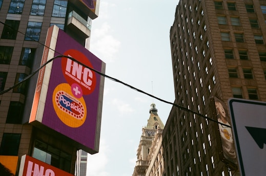 A cityscape featuring tall buildings with a prominent billboard displaying the logos for INC and Popsicle. A historical-style building with a detailed spire is visible in the background. The sky is partly cloudy, and a power line runs across the image.