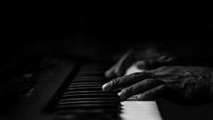 Hands are gently playing the keys of a piano or keyboard, casting soft shadows in the low light. A strong sense of focus and concentration is suggested by the close-up view.