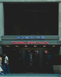 A marquee sign for The Tonight Show Starring Jimmy Fallon is displayed, featuring neon lights in blue and red. Two people are walking past the entrance, and the area is dimly lit with some visible posters or signage in the background.
