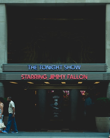 A marquee sign for The Tonight Show Starring Jimmy Fallon is displayed, featuring neon lights in blue and red. Two people are walking past the entrance, and the area is dimly lit with some visible posters or signage in the background.