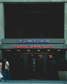 A marquee sign for The Tonight Show Starring Jimmy Fallon is displayed, featuring neon lights in blue and red. Two people are walking past the entrance, and the area is dimly lit with some visible posters or signage in the background.
