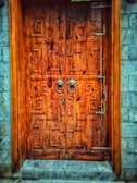 An intricately designed wooden door with geometric patterns carved into its surface. The door features a pair of ornate metal handles and is set within a stone wall structure. The wood has a rich, warm tone, and the stone wall contrasts with its cool gray hue.