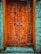 An intricately designed wooden door with geometric patterns carved into its surface. The door features a pair of ornate metal handles and is set within a stone wall structure. The wood has a rich, warm tone, and the stone wall contrasts with its cool gray hue.