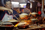 A person in a striped shirt and apron is cooking at a high-heat stove with several clay pots. Sparks fly around, creating a vibrant and dynamic scene.