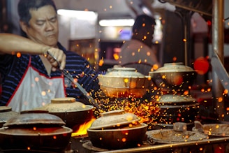 A person in a striped shirt and apron is cooking at a high-heat stove with several clay pots. Sparks fly around, creating a vibrant and dynamic scene.