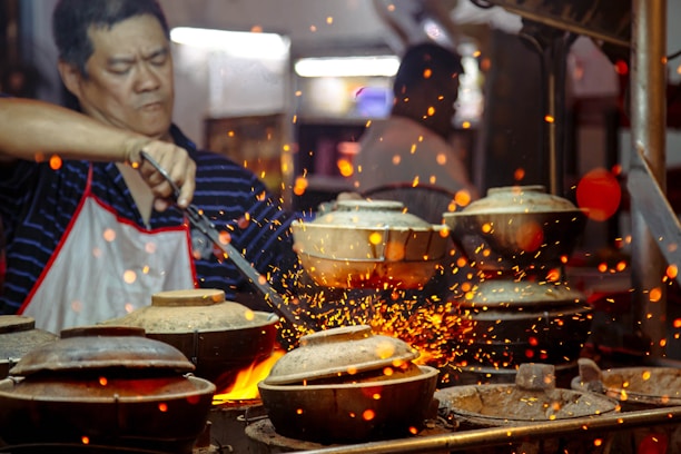 A person in a striped shirt and apron is cooking at a high-heat stove with several clay pots. Sparks fly around, creating a vibrant and dynamic scene.