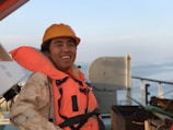 A person wearing an orange life jacket and hard hat is smiling on a ship's deck. The background includes a railing and the calm sea stretching into the horizon under a clear sky.
