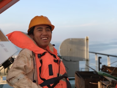 A person wearing an orange life jacket and hard hat is smiling on a ship's deck. The background includes a railing and the calm sea stretching into the horizon under a clear sky.