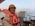 A person wearing an orange life jacket and hard hat is smiling on a ship's deck. The background includes a railing and the calm sea stretching into the horizon under a clear sky.