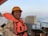 A person wearing an orange life jacket and hard hat is smiling on a ship's deck. The background includes a railing and the calm sea stretching into the horizon under a clear sky.
