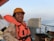 A person wearing an orange life jacket and hard hat is smiling on a ship's deck. The background includes a railing and the calm sea stretching into the horizon under a clear sky.