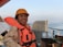 A person wearing an orange life jacket and hard hat is smiling on a ship's deck. The background includes a railing and the calm sea stretching into the horizon under a clear sky.