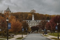 A picturesque campus scene with a central historic building featuring a bell tower, surrounded by autumn foliage in vibrant hues of orange and red. The foreground includes a wide pathway flanked by lampposts and banners. Cloudy skies create an overcast atmosphere.
