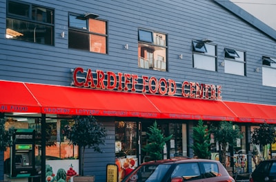 A storefront of a food center with a large red awning displaying the name 'Cardiff Food Centre' in bold lettering. The building has a modern design with multiple windows, and there are green plants in pots outside. Signs advertise various services offered, such as grocery, lottery, and fresh produce. A red car is parked in front of the entrance.