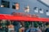 A storefront of a food center with a large red awning displaying the name 'Cardiff Food Centre' in bold lettering. The building has a modern design with multiple windows, and there are green plants in pots outside. Signs advertise various services offered, such as grocery, lottery, and fresh produce. A red car is parked in front of the entrance.