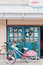 A pink bicycle is parked in front of a blue door decorated with colorful and artistic chalk drawings and signs indicating gluten-free and organic options. The door appears to be part of a shop or cafe. There are plants around the bicycle, creating a vibrant and inviting atmosphere.