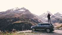 A person is standing on the roof of a dark-colored SUV, overlooking a mountainous landscape with snow-capped peaks. The scene conveys a sense of adventure and exploration with the vast, rugged terrain surrounding the vehicle.