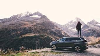 A person is standing on the roof of a dark-colored SUV, overlooking a mountainous landscape with snow-capped peaks. The scene conveys a sense of adventure and exploration with the vast, rugged terrain surrounding the vehicle.
