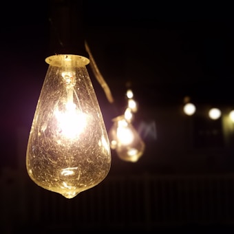 A close-up view of a glowing string light bulb with a warm, yellowish hue. The bulb is the main focus, with intricate details of the glass filament visible. Several blurred bulbs are seen in the background against a dark setting, creating a cozy atmosphere.
