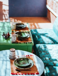 Two tables set for dining with ornate ceramic plates and cups. The tables have green and round surfaces, with one table also featuring a green sofa with patterned cushions. The setup includes small condiment containers, suggesting a cozy and intimate dining atmosphere. Sunlight filters through creating shadows and highlights on the scene.
