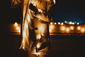 A person is dressed in a cozy beige coat with warm string lights wrapped around them. The background features a softly blurred cityscape with additional string lights creating a warm and festive ambiance.
