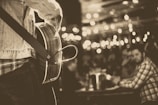 A musician is playing an acoustic guitar on stage, wearing a denim jacket. The focus is on the guitar and the musician's upper body. In the background, out of focus, an audience is seated at tables with drinks. Ambient string lights create a warm, intimate atmosphere.