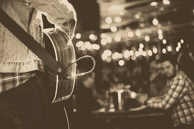 A musician is playing an acoustic guitar on stage, wearing a denim jacket. The focus is on the guitar and the musician's upper body. In the background, out of focus, an audience is seated at tables with drinks. Ambient string lights create a warm, intimate atmosphere.