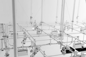 A complex arrangement of white metal pipes and fixtures suspended from the ceiling, creating an intricate network of lines and angles. The setup includes various mechanical components and technical devices, contributing to an industrial and architectural aesthetic.