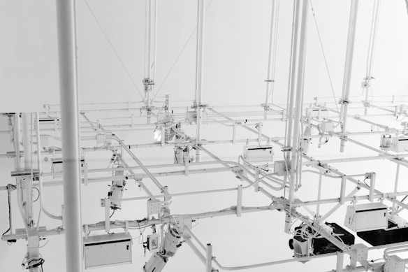 A complex arrangement of white metal pipes and fixtures suspended from the ceiling, creating an intricate network of lines and angles. The setup includes various mechanical components and technical devices, contributing to an industrial and architectural aesthetic.