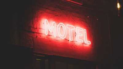 A glowing red neon sign displaying the word 'MOTEL' is mounted on a brick wall. The brick texture is visible in the dimly lit atmosphere, with the red neon light casting a soft glow around the sign.