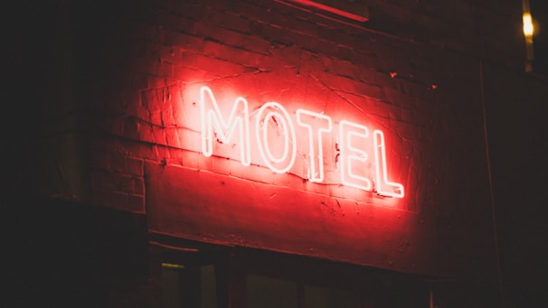 A glowing red neon sign displaying the word 'MOTEL' is mounted on a brick wall. The brick texture is visible in the dimly lit atmosphere, with the red neon light casting a soft glow around the sign.