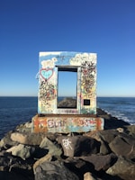 A wooden frame covered in colorful graffiti stands on a rocky jetty by the sea. The frame has various drawings and writings, including a heart with initials and other abstract designs. The ocean stretches out under a clear blue sky.