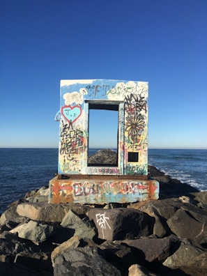 A wooden frame covered in colorful graffiti stands on a rocky jetty by the sea. The frame has various drawings and writings, including a heart with initials and other abstract designs. The ocean stretches out under a clear blue sky.