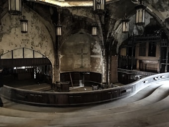 An abandoned, dimly lit church interior with worn and peeling walls, showcasing gothic architectural elements. The large space features arching ceilings and a prominent cross on a central wall. Ornate, hanging lanterns provide minimal lighting. The wooden pews and balconies are visible, reflecting the church's state of disrepair yet retaining some grandeur.