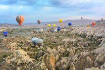 Numerous colorful hot air balloons float over a dramatic landscape with unique rock formations and valleys. The sky is mostly clear with a few clouds, and the scene conveys a sense of adventure and serenity.