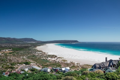 A panoramic view of a pristine white sandy beach bordered by turquoise ocean waters. The coastline curves gently, leading to verdant hills in the background. Foreground features include a cluster of houses with distinct thatched roofs amidst lush greenery, adding a touch of architectural interest. A range of mountains can be seen in the distance under a clear blue sky.