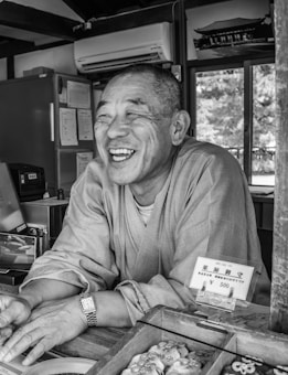 A man is smiling warmly while leaning on a counter with various items displayed in front. The setting appears to be indoors, with an air conditioner mounted on the wall and a framed picture of a traditional building. Natural light filters through a window, partially visible in the background.