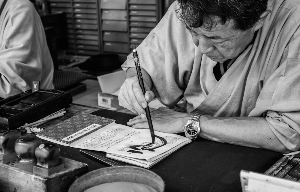 grayscale photo of man sitting on chair in front of table while writing