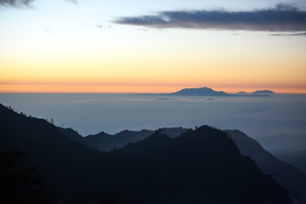 The image captures a serene landscape with silhouetted mountains in the foreground under a pastel-toned sky at dawn or dusk. The sky showcases a gradient from pale orange to light blue, with wispy clouds above a layer of mist that envelops distant mountain peaks.