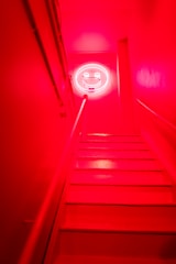 A staircase with bright red walls and steps leading up to a neon smiley face sign on the wall at the top. The lighting creates an intense and vibrant atmosphere.