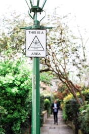 A green lamp post with a 'Neighbourhood Watch' sign is prominently placed in a lush garden setting with trees and bushes. In the background, a person is walking along a path lined with greenery and flowers.