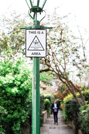 A green lamp post with a 'Neighbourhood Watch' sign is prominently placed in a lush garden setting with trees and bushes. In the background, a person is walking along a path lined with greenery and flowers.