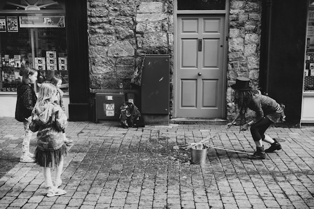 A street performer wearing a top hat and casual clothes is interacting with a group of children. The performer is squatting and using a tool to handle a bucket placed on a cobblestone street. The scene is set in front of a brick wall and a closed door, creating an urban backdrop.